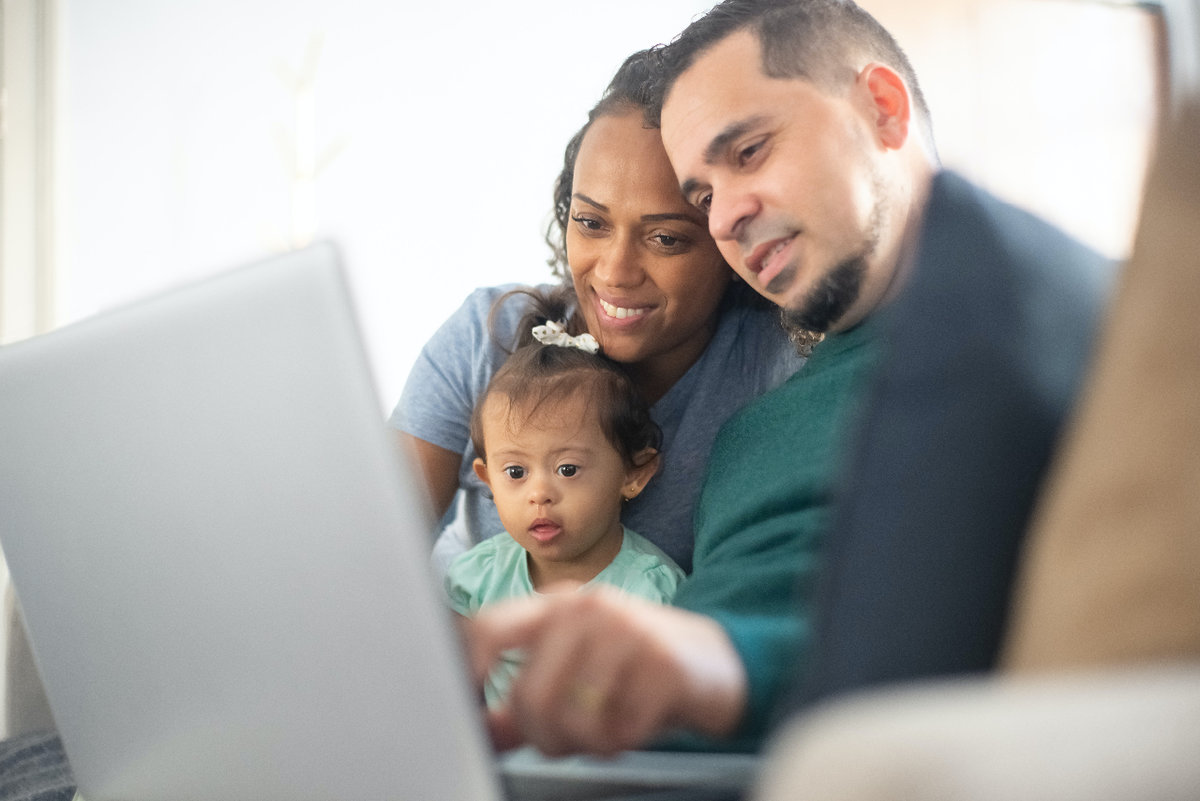 A young family (mom, dad, infant girl) sit together and look at something on a laptop together.